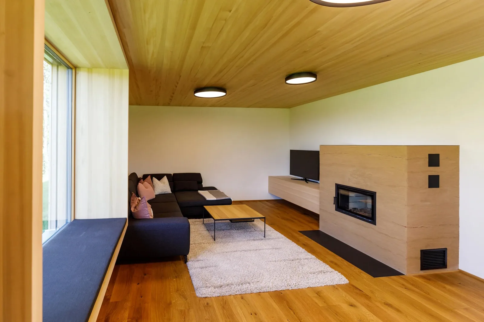 Wohnraum in Holz mit einem Ofen in heller Travertin-Verkleidung und breiter Sichtscheibe