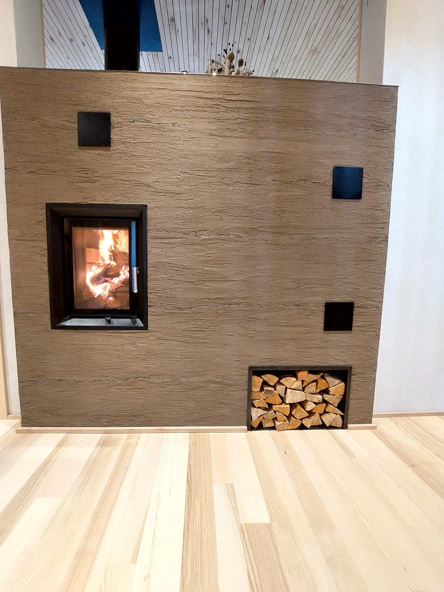 Kamin mit Sichtscheibe in einer gespachtelten Lehmputzwand, daneben ein Fach mit Scheitholz